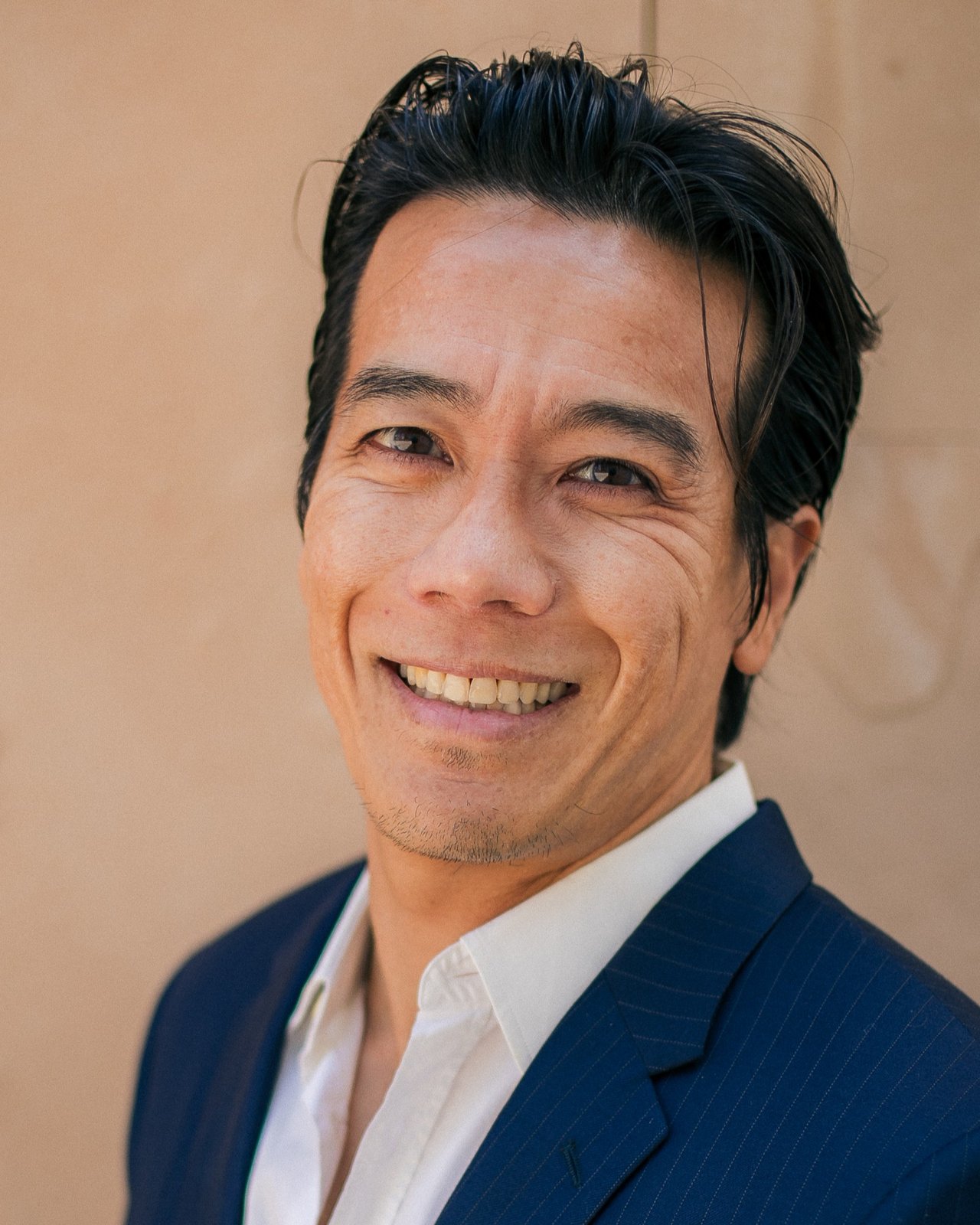 Patrick P. Phancao, smiling, in a navy suit and open white shirt, photographed against a sandstone wall.