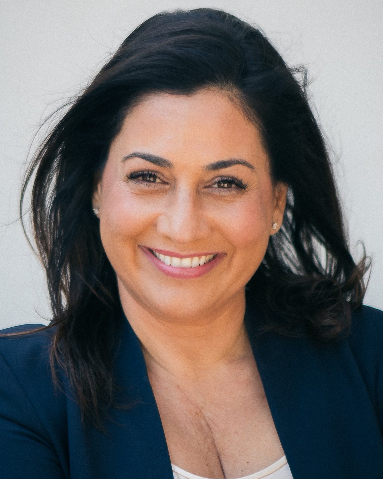 Shadi Ala’i Shaffer, smiling, in a navy jacket, photographed against a pale wall.